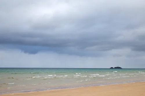 This landscape photograph depicts Godrevy beach in Cornwall, England, during the early evening in summer. The scene highlights the natural colours of the beach under a cloudy sky, with gentle waves washing onto the sandy shore and the turquoise sea stretching out towards the horizon. To the right, the image features Godrevy Island with its recognisable lighthouse, a notable landmark on this part of the Cornish coast. The overall atmosphere is characteristic of the United Kingdom’s coastal scenery, with muted tones and a wide expanse of open water typical of a summer evening by the sea.